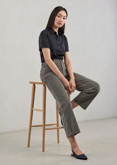 A woman wearing a black short sleeve polo shirt sits on a wooden stool, paired with olive green trousers and black flats.