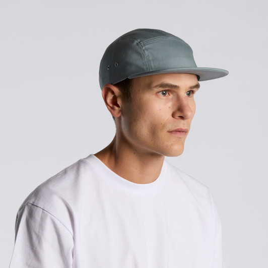 A five-panel cap in a soft blue-grey colour worn by a model, featuring a subtle logo.