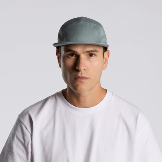 A five-panel cap in grey being worn by a model with a plain white t-shirt, featuring a logo on the front.