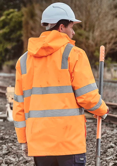 A person wearing an orange waterproof jacket with reflective stripes and a white hard hat, standing near railway tracks.