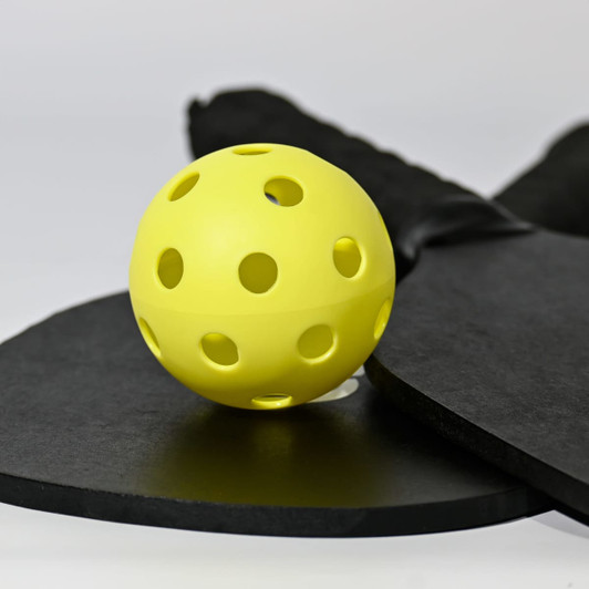 A yellow pickleball with holes, resting on a black paddle and a round black surface, featuring a logo.