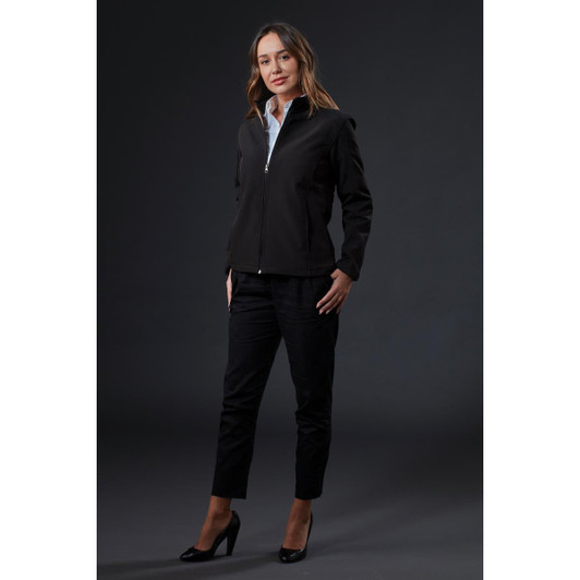 A black softshell jacket is worn by a woman, styled with black pants and heels, against a dark background.