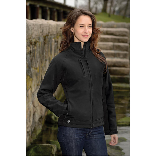 A woman stands by a stone pathway wearing a black soft shell jacket, featuring a zippered pocket and a logo.
