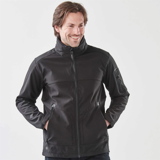 A dark black softshell jacket with a full zip, featuring side pockets and a collar, worn by a smiling man.