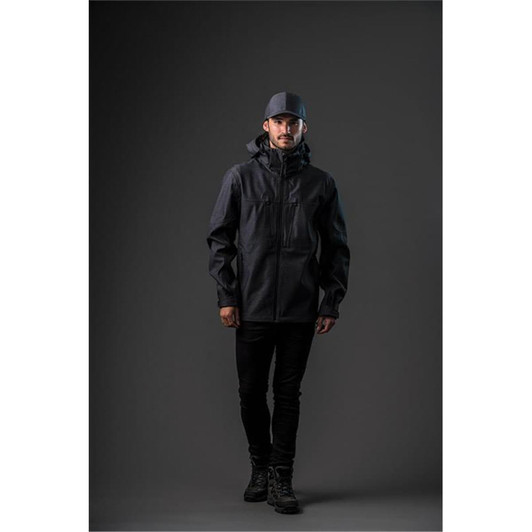 A man wearing a black softshell jacket and matching cap, standing against a dark background. The jacket has a logo.