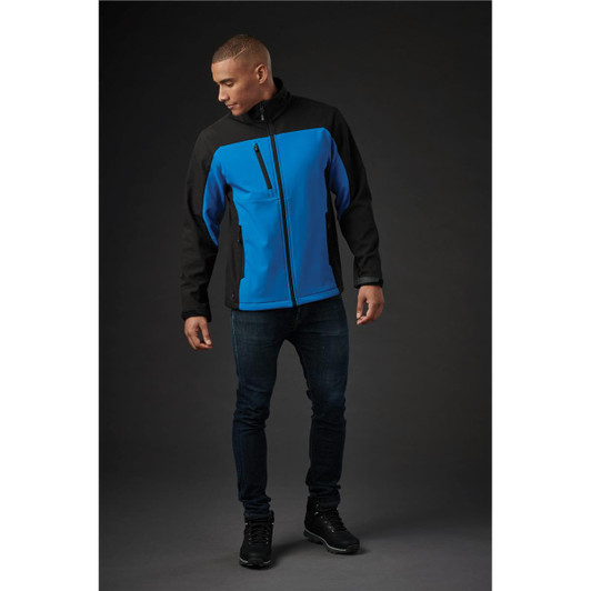 A men's softshell jacket in blue and black, featuring a zippered pocket and a logo. Styled with dark jeans and shoes.