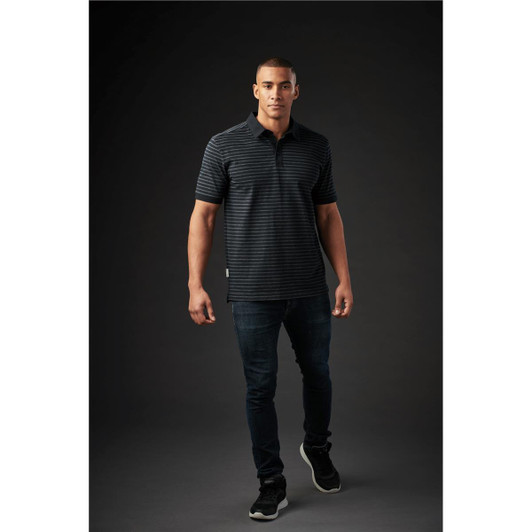 A man models a black and dark grey striped polo shirt, paired with dark jeans and sneakers. The shirt has a logo.