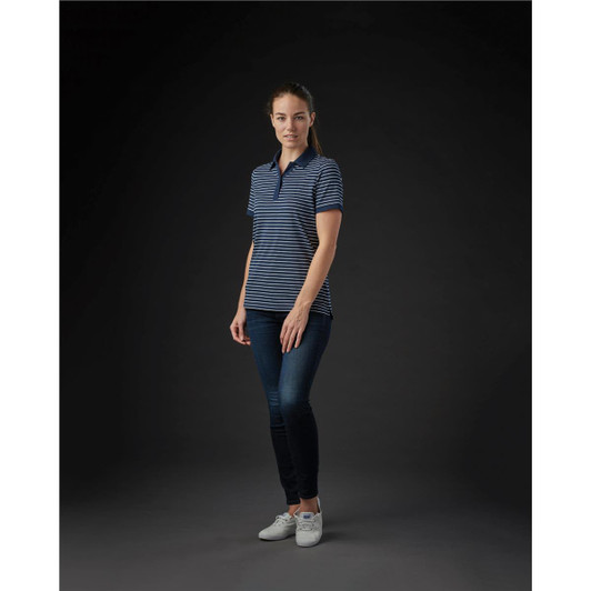 A woman wearing a navy and white striped polo shirt, paired with blue jeans and white shoes, standing against a dark background.