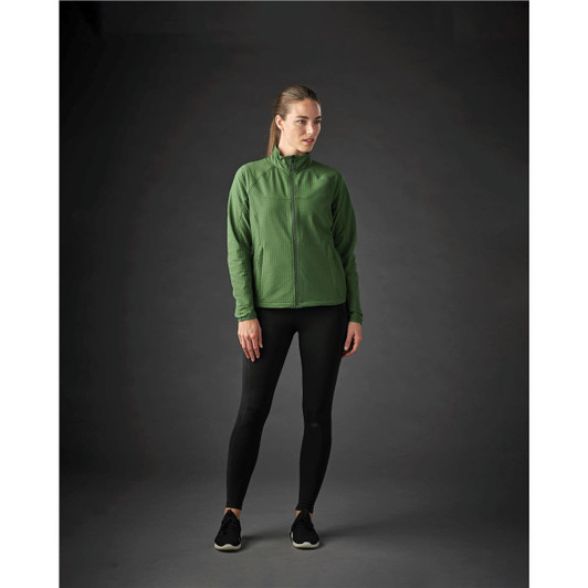 A woman stands wearing a green Women’s Kyoto Jacket paired with black leggings and shoes, with a dark background.