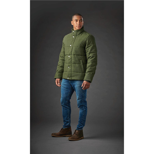 A man stands wearing a green thermal jacket with a buttoned front, paired with jeans and boots. The background is grey.