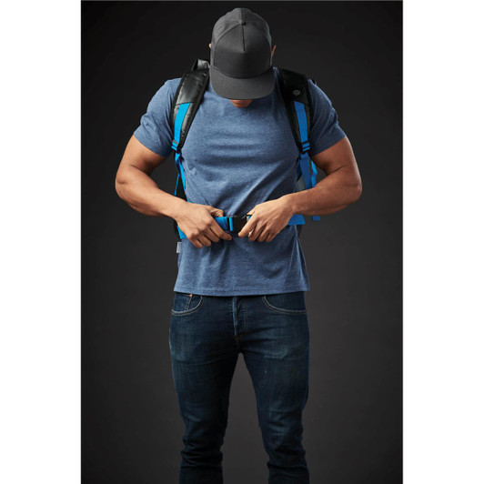 A person adjusts the straps on a blue Kemano Backpack while wearing a grey T-shirt and a cap. The backpack features black accents.