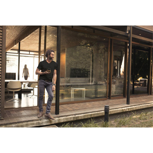 A man wearing a black V-neck tee stands outside a modern building with large windows. The setting features greenery.