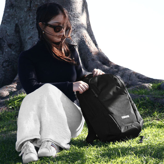A black Thule Recycled Lumion 16" computer backpack sits on the grass beside a person wearing sunglasses.