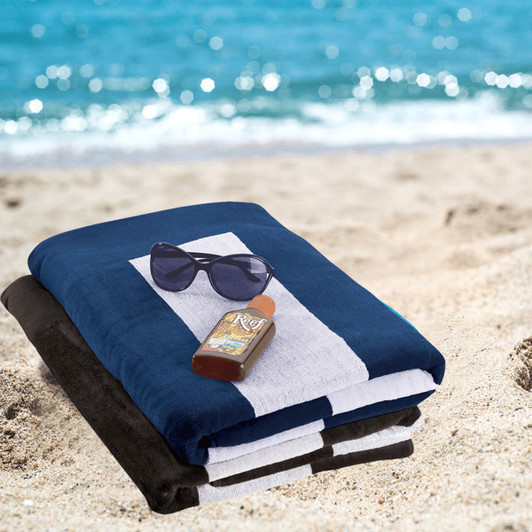 A stack of beach towels in navy blue and black, with sunglasses and sunscreen, placed on sandy beach.