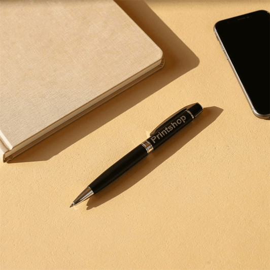 A silver ballpoint pen rests beside a notebook and a smartphone on a beige surface. The pen has a logo.