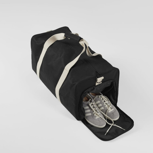 A black duffle bag featuring a white strap, with a pair of grey shoes visible inside. The bag has a logo.
