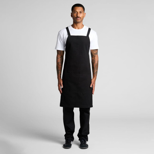 A black apron with a bib design worn over a white T-shirt by a model, featuring shoulder straps and a logo.