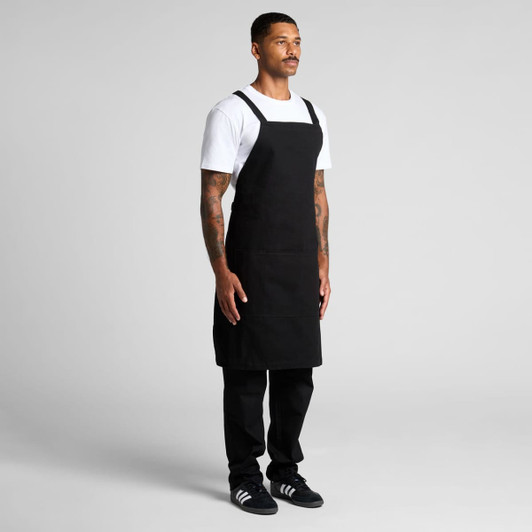 A black Carrie apron, featuring adjustable straps, displayed on a model in a standing pose against a plain background.