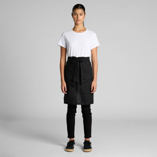 A linen half apron in black is worn over a white t-shirt and black trousers, featuring a tied waist.