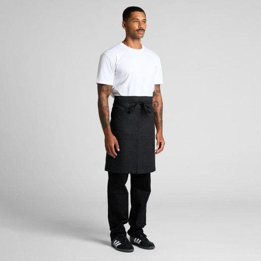 A man wearing a black linen half apron over a white t-shirt and black pants, standing confidently.
