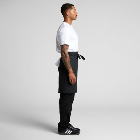 A man stands sideways wearing a black linen half apron over black pants and a white t-shirt.