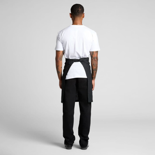 A black linen half apron is worn by a person facing away, paired with a white t-shirt and black trousers.