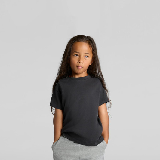 A child wearing a black heavy faded tee, with long hair and hands in pockets, set against a neutral background.