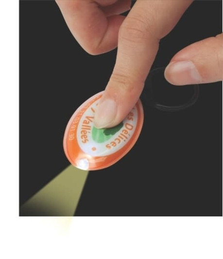 A hand pressing an orange press-light keyring, emitting light. The keyring features a logo on its surface.
