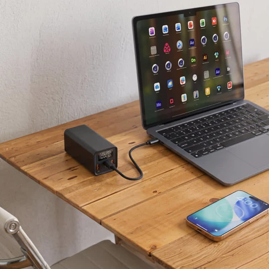 A black power bank is connected to a laptop on a wooden table, with a smartphone nearby.