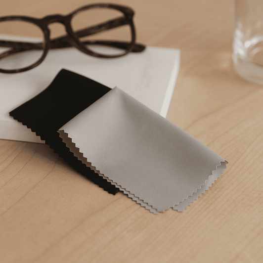 A grey and black mini microfibre cleaning cloth sits on a wooden surface beside glasses and a glass of water.