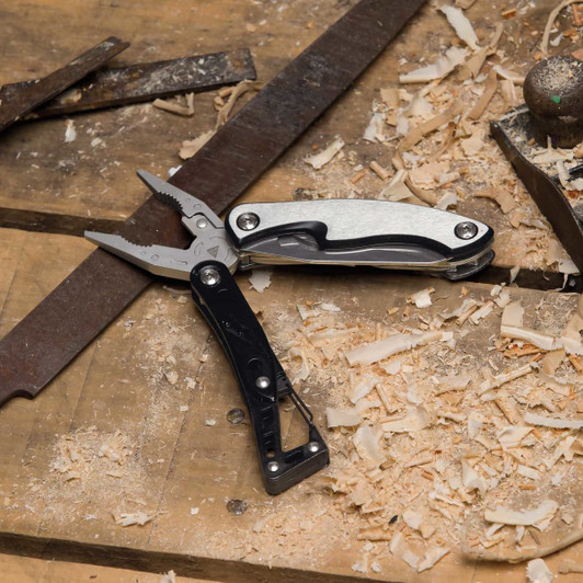 A multi-tool with black and silver accents, featuring pliers and various tools, placed on a wooden surface with shavings.