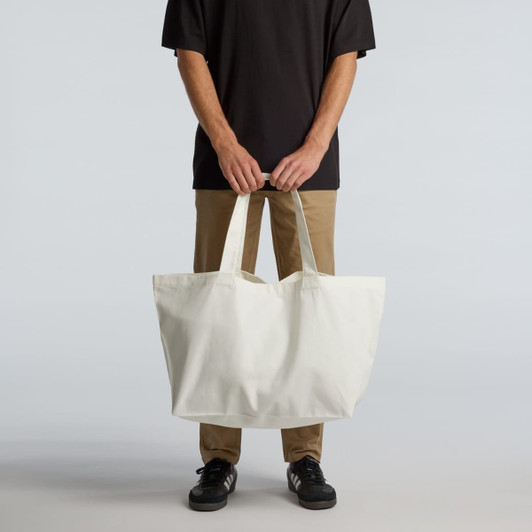 An oversized tote bag in off-white held by a person, featuring strong handles and a simple design.