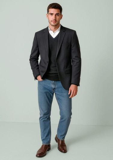 A man in a dark blazer over a black sweater, wearing light blue jeans and brown shoes, stands against a pale background.
