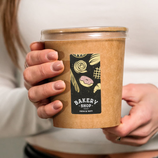 A hand holding a matte rectangular sticker on a brown takeout container, featuring bakery-themed illustrations.