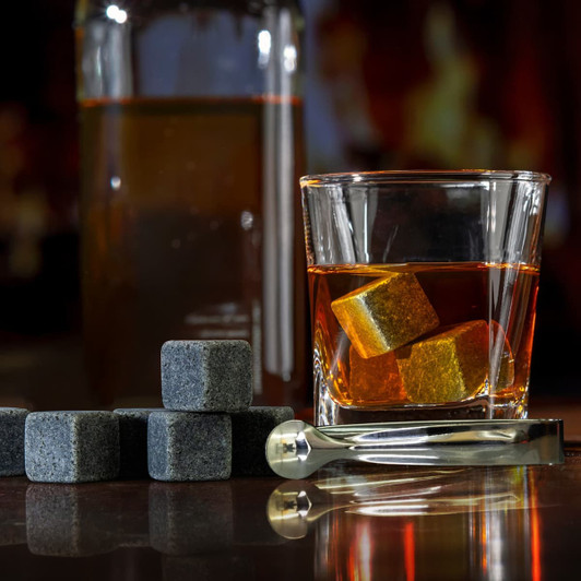 A glass with whiskey and grey whiskey stones beside a drink bottle and a stainless steel tongs.