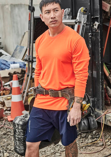 An orange long sleeve safety tee is worn by a man with a tool belt, set against a cluttered worksite background.