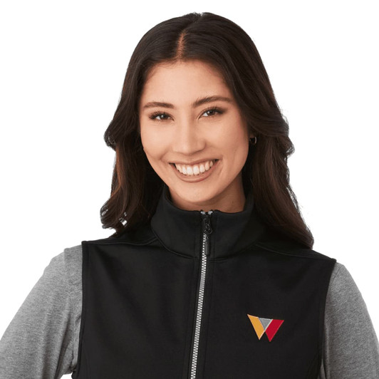 A black softshell vest featuring a zippered front and a logo. The model wears a grey long-sleeve top underneath.