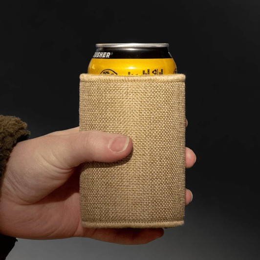 A jute stubby holder in a natural beige colour, holding a can, being held by a hand.