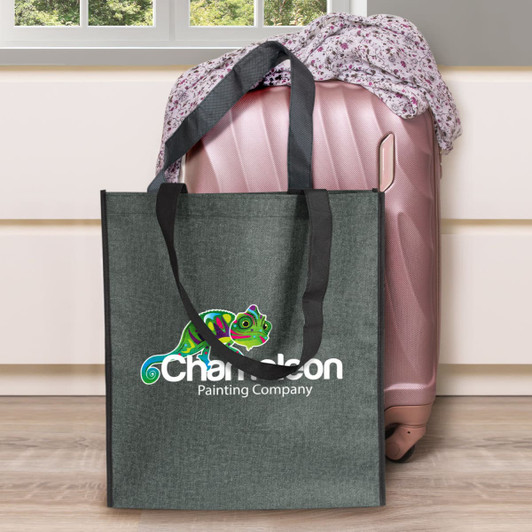 A grey tote bag with black handles, featuring a logo. Positioned beside a pink suitcase and patterned fabric.