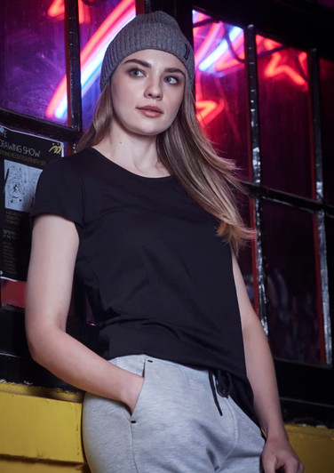 A woman in a black t-shirt and grey pants leans against a window, wearing a grey beanie. The background features neon lights.