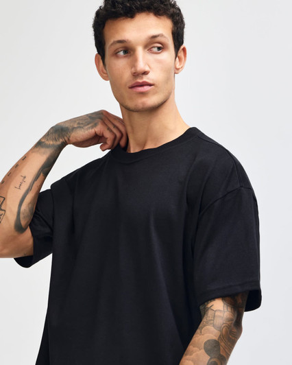 A model wearing a black American Apparel Super Heavyweight T-Shirt, featuring a relaxed fit and visible tattoos.