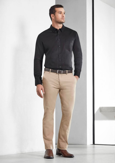 A man wearing a black long sleeve shirt paired with beige trousers and brown shoes, standing against a plain background.