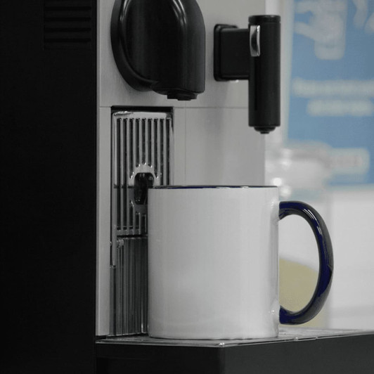 A two-tone ceramic mug with a white body and blue handle sits under a coffee machine. It has a logo on the side.