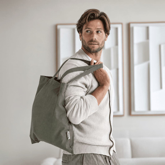 A man carries a large grey tote bag, wearing a light sweater, in a modern interior setting. The bag has a logo.