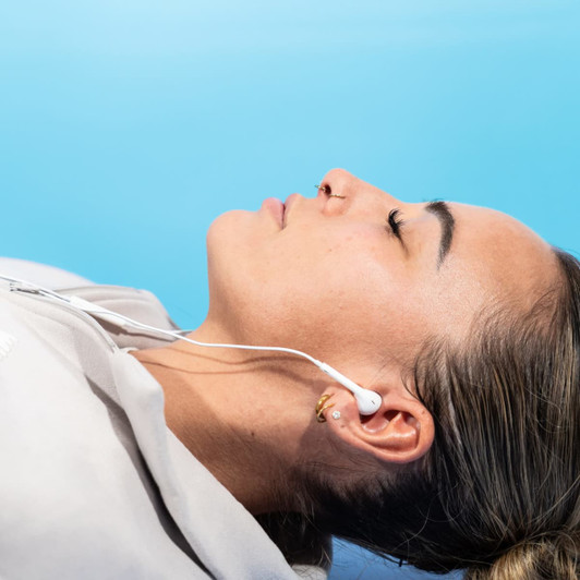 Earphones with a white cable connected to a user lying down against a blue background. A logo is visible on the earphones.