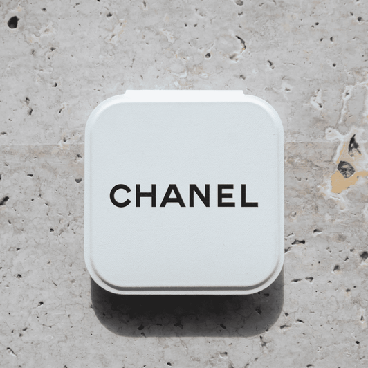 A compact mirror in a white case with a logo on the top, set against a textured stone background.