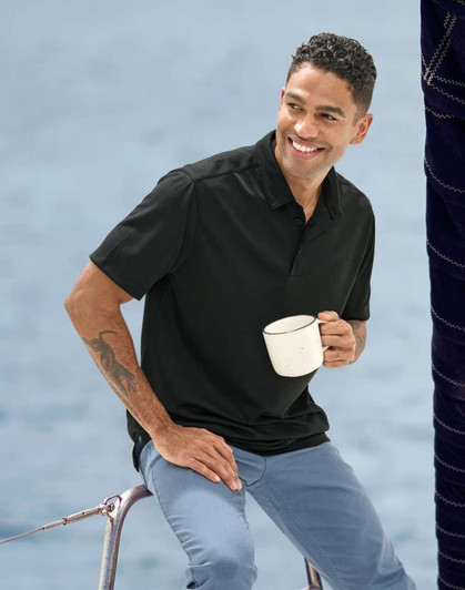 A man sits on a railing, wearing a black sustainable jacquard knit polo shirt and holding a white mug.