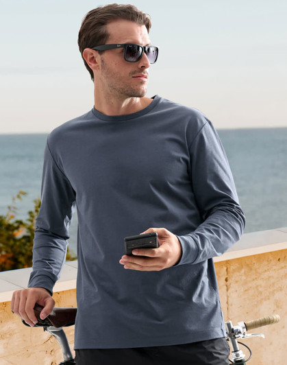 A man wearing a dark blue long-sleeve cotton tee shirt, holding a phone beside a bicycle with a coastal background.