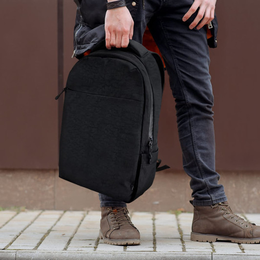 A black daily backpack is being held by a person wearing dark jeans and brown boots, standing on a paved surface.