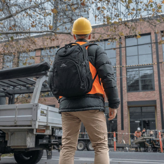 A black backpack worn by a person in an orange safety vest and yellow hard hat, standing outdoors.